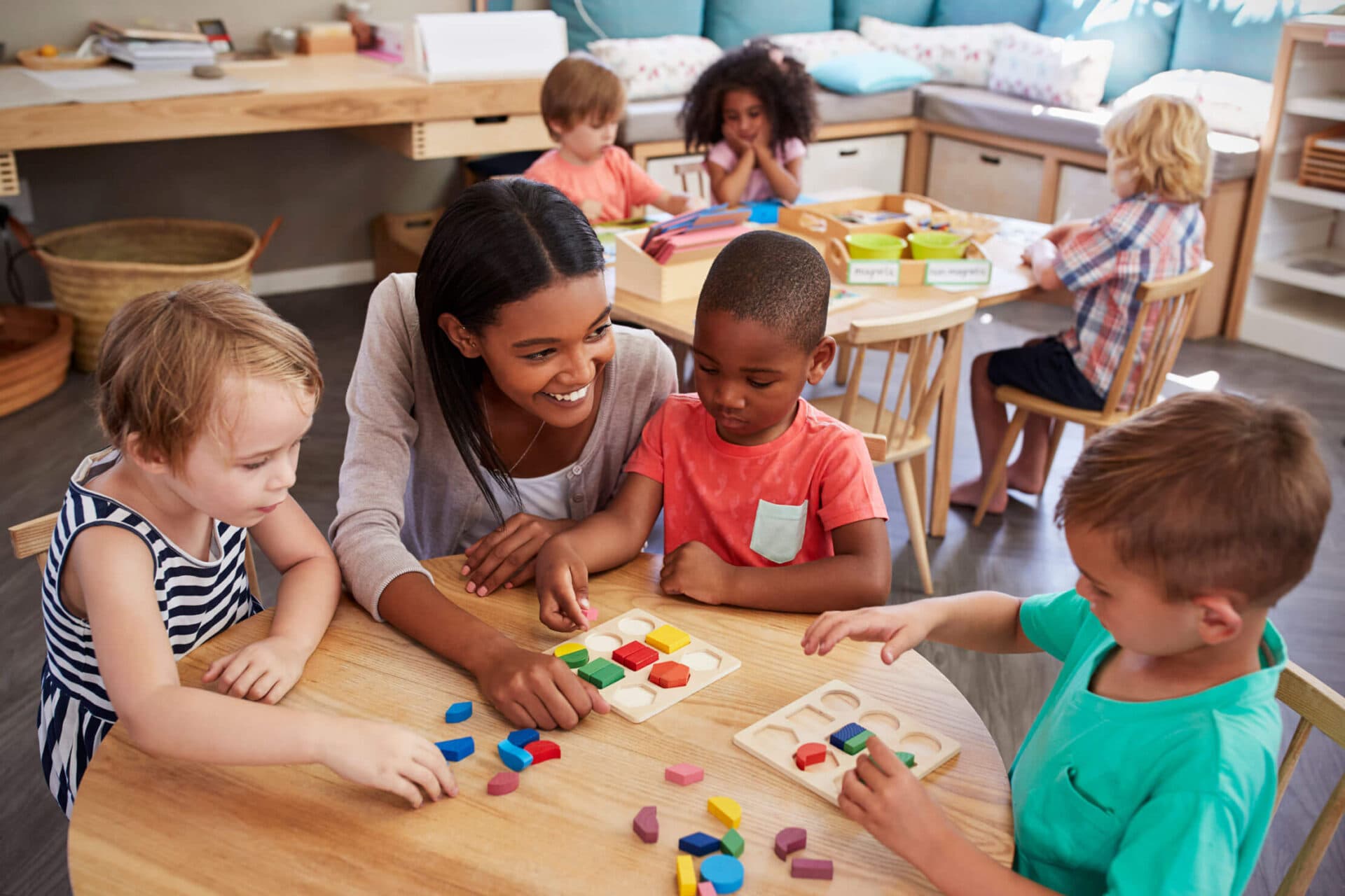 Teacher seated at a classroom table guiding four young children through a wooden shape-sorting activity