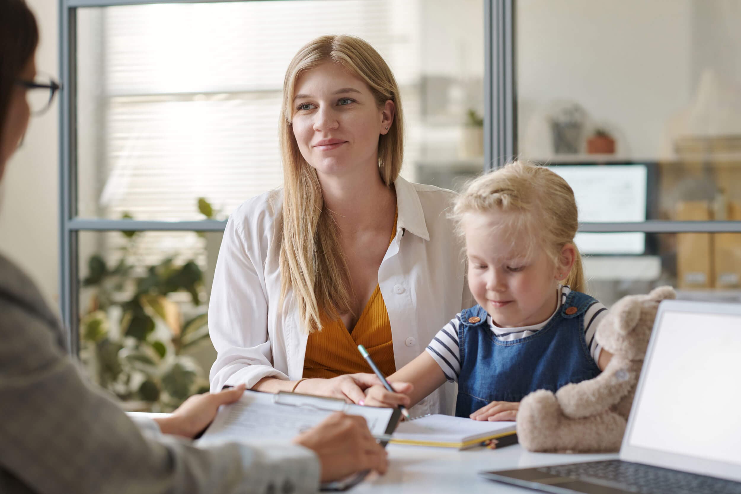 Parent and her young daughter meeting with a teacher who is reviewing notes on a clipboard